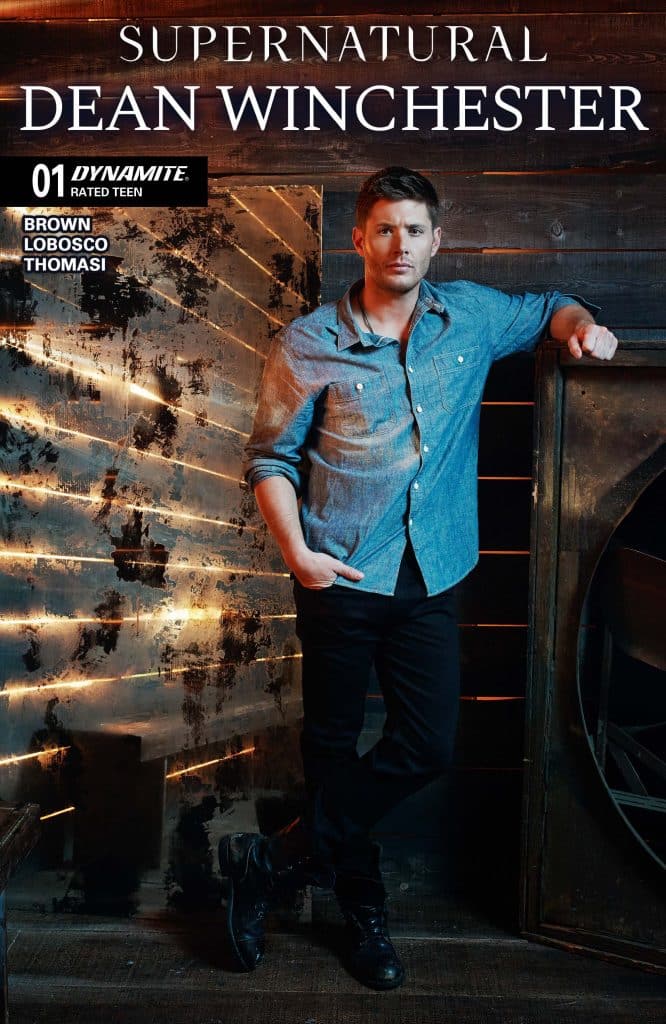 Promotional image of Dean Winchester from the TV series Supernatural, featuring him standing against a rustic backdrop with wooden elements. He wears a denim shirt and black pants.