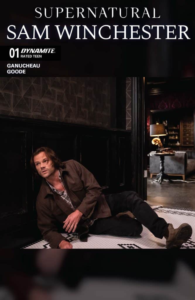 A character from Supernatural, Sam Winchester, is crouching on the floor in a dimly lit room, wearing a brown jacket and a plaid shirt.